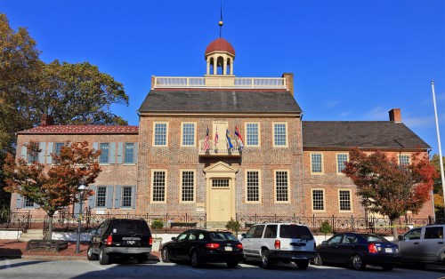 Historic building with a cupola, flags, and autumn trees, surrounded by parked cars under a clear blue sky.