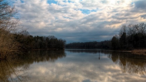 A serene lake reflects cloudy skies and surrounding trees, creating a tranquil natural scene.