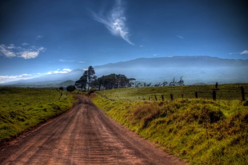 A dirt road winds through lush green fields, leading towards distant mountains under a clear blue sky.