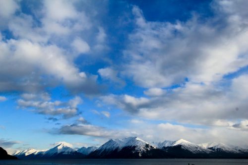Snow-capped mountains under a blue sky with scattered clouds, reflecting on a calm body of water.