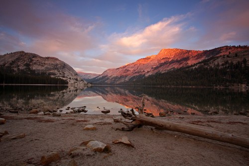A serene lake at sunset, reflecting mountains and colorful clouds, with a log and rocks in the foreground.