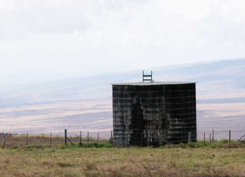 A weathered cylindrical water tank on a grassy landscape with distant hills under a cloudy sky.