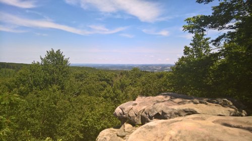 A scenic view from a rocky outcrop, overlooking lush green trees and distant hills under a blue sky with clouds.