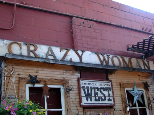 Sign on a rustic building reads "CRAZY WOMAN" with decorative stars and flowers below.