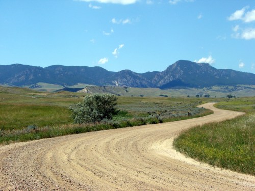 A winding dirt road leads through green fields, with mountains in the background under a clear blue sky.