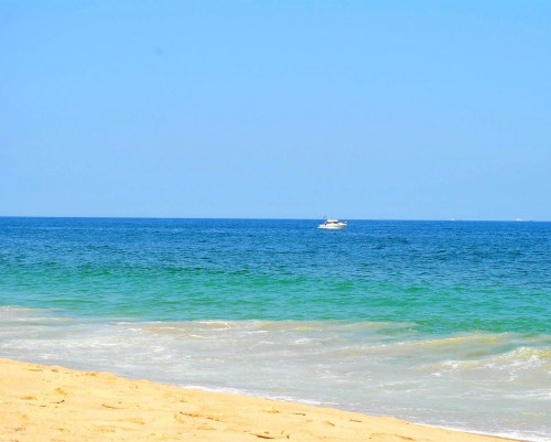 A serene beach scene with golden sand, gentle waves, and a small boat on the calm blue ocean under a clear sky.