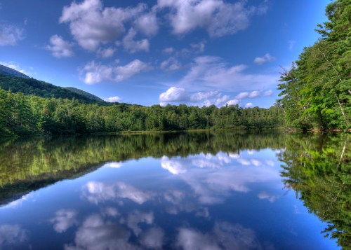 A serene lake reflecting blue skies and clouds, surrounded by lush green trees and mountains in the background.