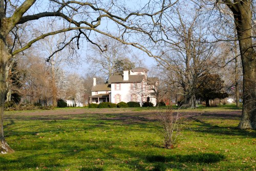 A large, historic house surrounded by trees and a grassy area, with a clear blue sky in the background.