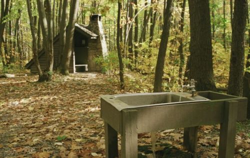A rustic outdoor sink in a forest, with a small cabin visible in the background among colorful autumn leaves.