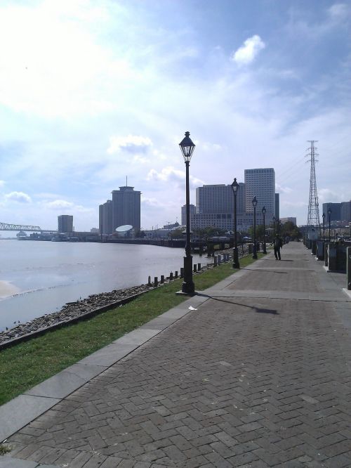 A waterfront walkway lined with lampposts, leading to city buildings under a bright sky.