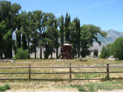 A red wooden structure surrounded by tall trees and grassy fields, with mountains in the background under a clear blue sky.