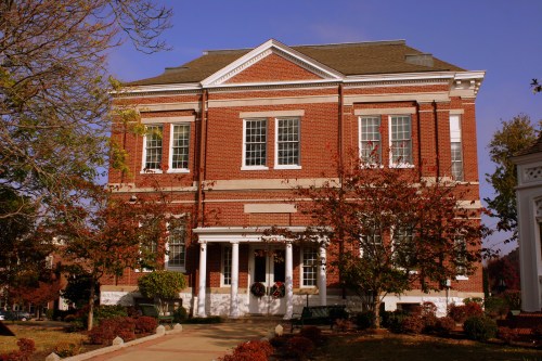A brick building with white trim, featuring large windows and a porch, surrounded by autumn foliage.