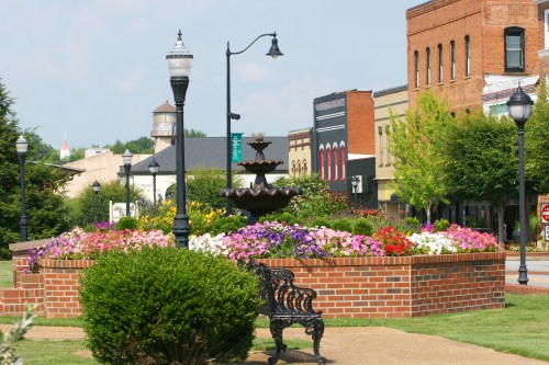 A vibrant flower bed surrounds a fountain in a town square, with brick buildings and lampposts in the background.