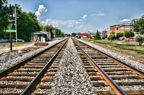 A view of railway tracks stretching into the distance, flanked by greenery and buildings under a blue sky.