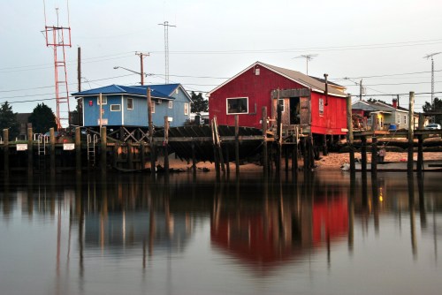 Colorful houses on stilts by a calm waterway, reflecting in the water at dusk. Power lines in the background.