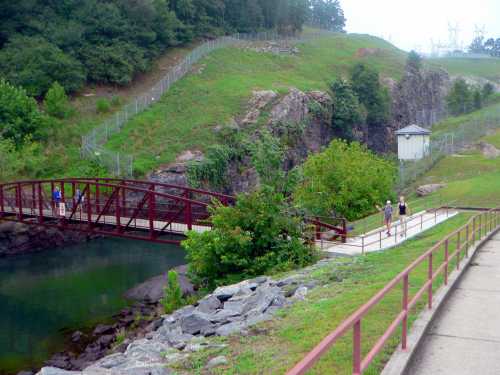 A scenic view of a walking path with a red bridge over a calm waterway, surrounded by greenery and rocky terrain.