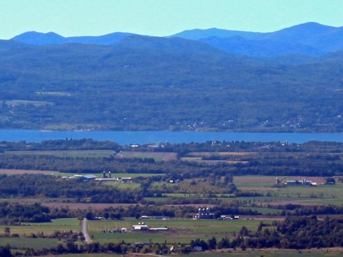 A scenic view of rolling hills, farmland, and a lake surrounded by mountains under a clear blue sky.