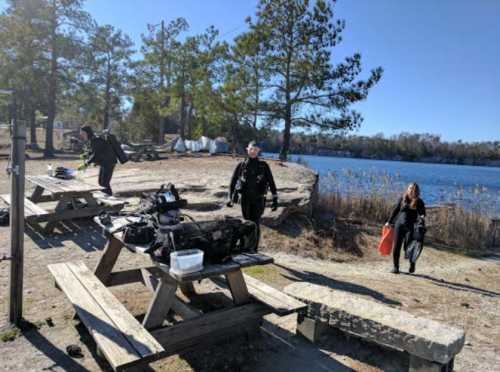 Two people near a lake, one carrying a bag, the other preparing equipment on a picnic table surrounded by trees.