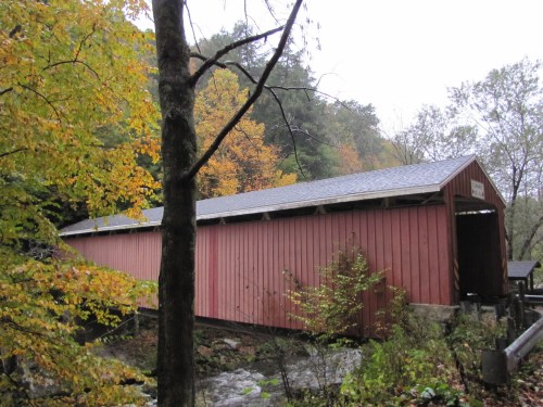 A red covered bridge surrounded by autumn trees and a stream, set in a misty, serene landscape.