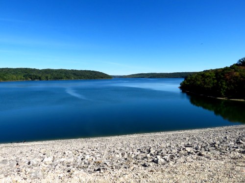 A serene lake surrounded by lush greenery under a clear blue sky, with a rocky shoreline in the foreground.