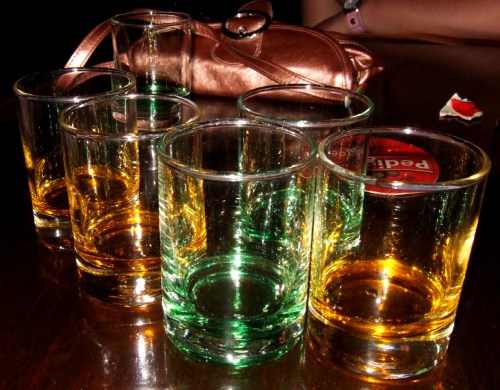 A collection of empty glass tumblers in green and amber colors on a wooden table, with a small purse in the background.