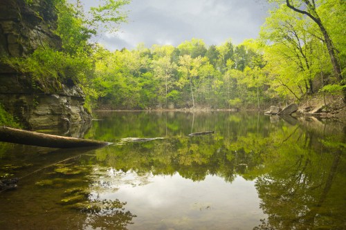 A serene lake surrounded by lush green trees and rocky cliffs, reflecting the cloudy sky above.