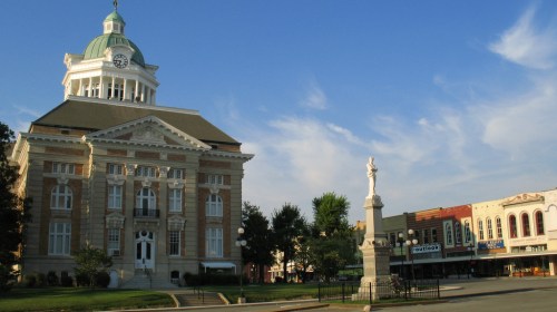 Historic courthouse with a clock tower, surrounded by trees and a monument, against a clear blue sky.