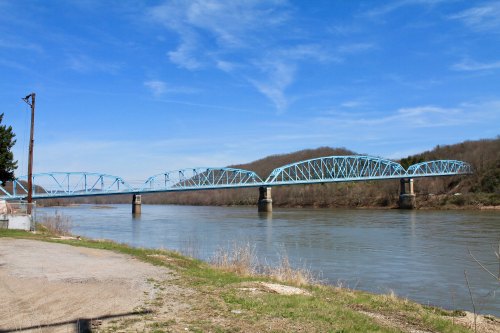 A blue arch bridge spans a river, surrounded by green hills under a clear blue sky.