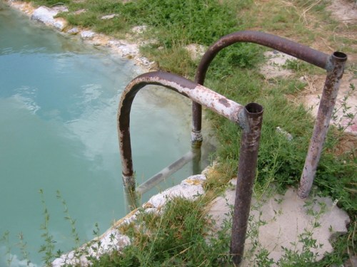 Rusty metal handrails beside a turquoise pool, surrounded by overgrown grass and a rocky edge.