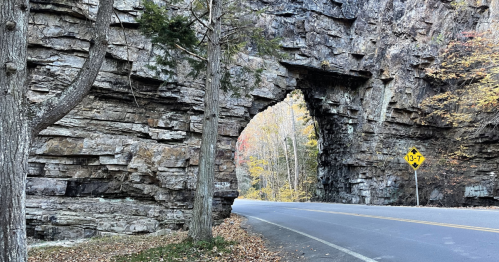 A stone archway in a rocky landscape, with trees nearby and a road sign indicating a height limit. Autumn foliage visible.