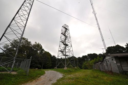 Two tall communication towers stand near a gravel path, surrounded by trees and a fenced area under a cloudy sky.