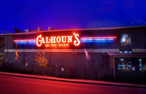 Neon sign of Calhoun's on the River restaurant illuminated at dusk, with a rustic building exterior.