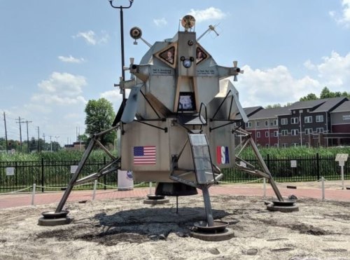 A replica of a lunar lander with the American flag, set in a park against a blue sky and buildings in the background.