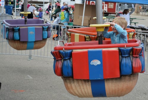 A child plays in a colorful amusement park ride, surrounded by other attractions and people in the background.
