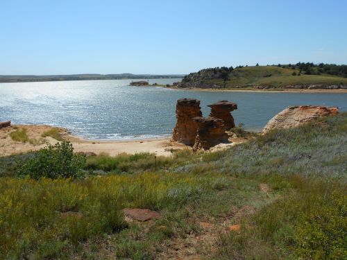 Scenic view of a calm lake with rocky formations and green hills under a clear blue sky.