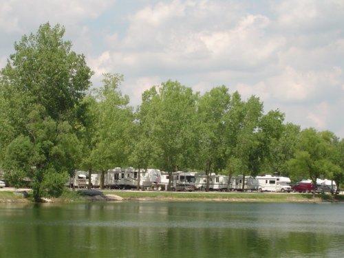 A serene lakeside scene with RVs parked under lush green trees and a cloudy sky in the background.
