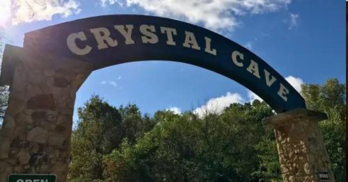 Sign for Crystal Cave with a blue arch and stone pillars, surrounded by trees under a partly cloudy sky.