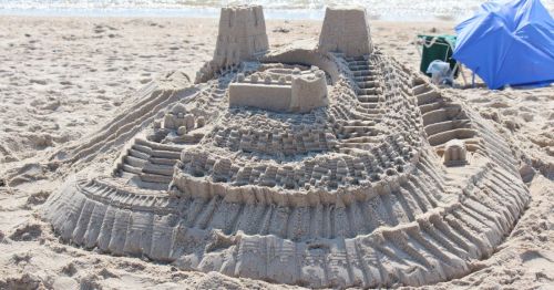 Intricate sandcastle with towers, steps, and detailed patterns on a beach, with ocean waves in the background.