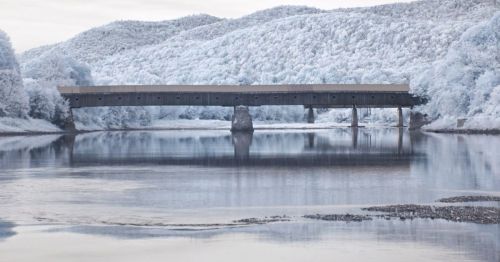 A serene landscape featuring a bridge over a calm river, surrounded by trees and mountains in a soft, muted color palette.