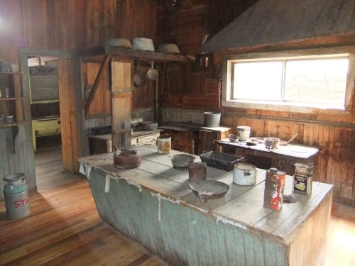 A rustic kitchen with wooden walls, a large table, and various pots and containers scattered around.