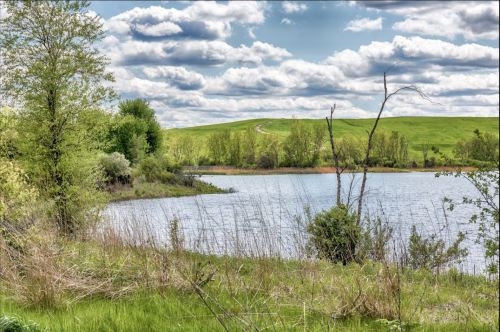 A serene lakeside scene with lush greenery, gentle hills, and a cloudy sky reflecting on the water's surface.