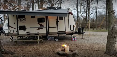 A camper parked under trees, with a picnic table nearby and a small fire pit in front, creating a cozy outdoor scene.