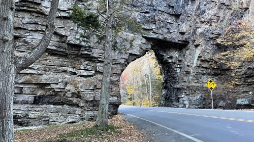 A stone archway in a rocky cliff beside a road, surrounded by trees and autumn foliage. A road sign is visible.
