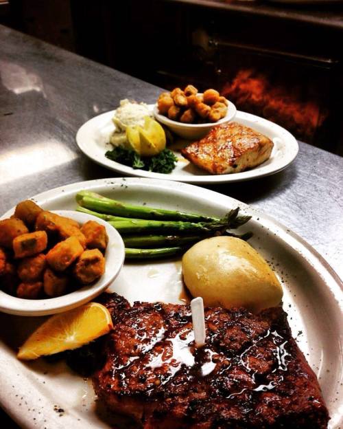 A plate of grilled steak with a roll, okra, asparagus, and lemon, alongside a second plate of shrimp and fish.