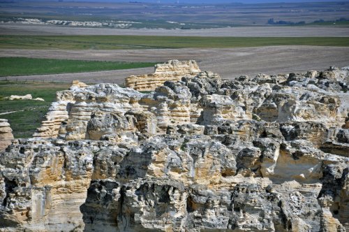 A panoramic view of rugged, layered rock formations against a backdrop of green fields and distant plains.
