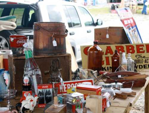 A table filled with vintage items, including bottles, signs, and wooden boxes, at an outdoor market.