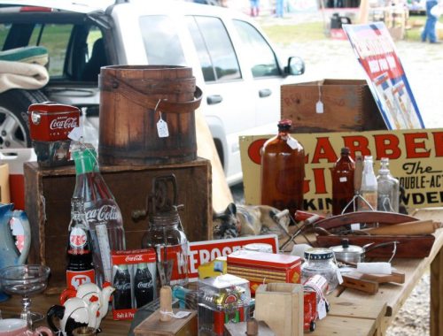 A table filled with vintage items, including bottles, signs, and wooden boxes, at an outdoor market.