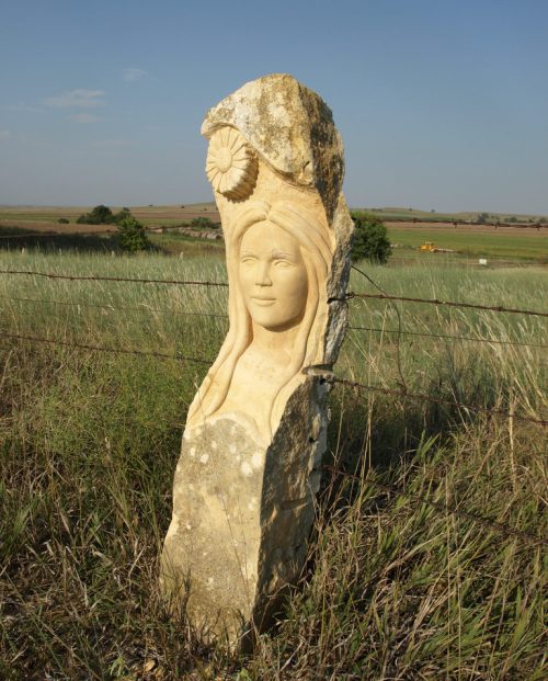 A carved stone sculpture of a woman's face, set against a grassy landscape under a clear blue sky.