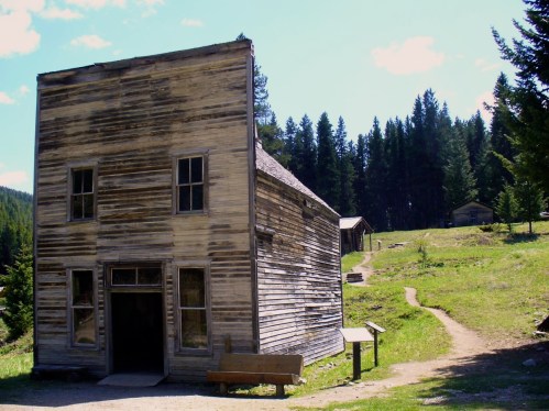 A weathered wooden building in a forested area, with a path leading to other structures in the background.