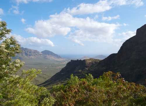 A scenic view of mountains and valleys under a partly cloudy sky, with the ocean visible in the distance.